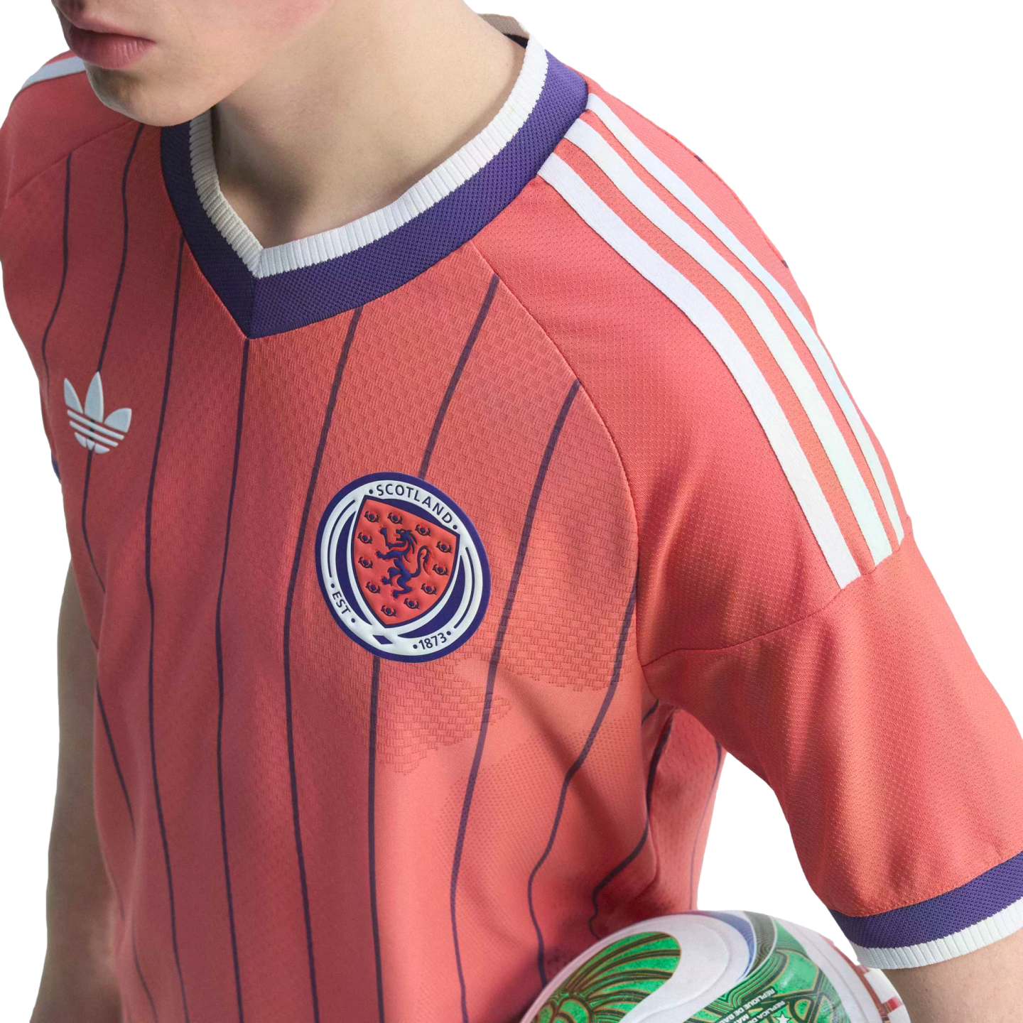 A person wears a pink Scotland national football team jersey with purple pinstripes, white Adidas shoulder stripes, and the team crest while holding a soccer ball.