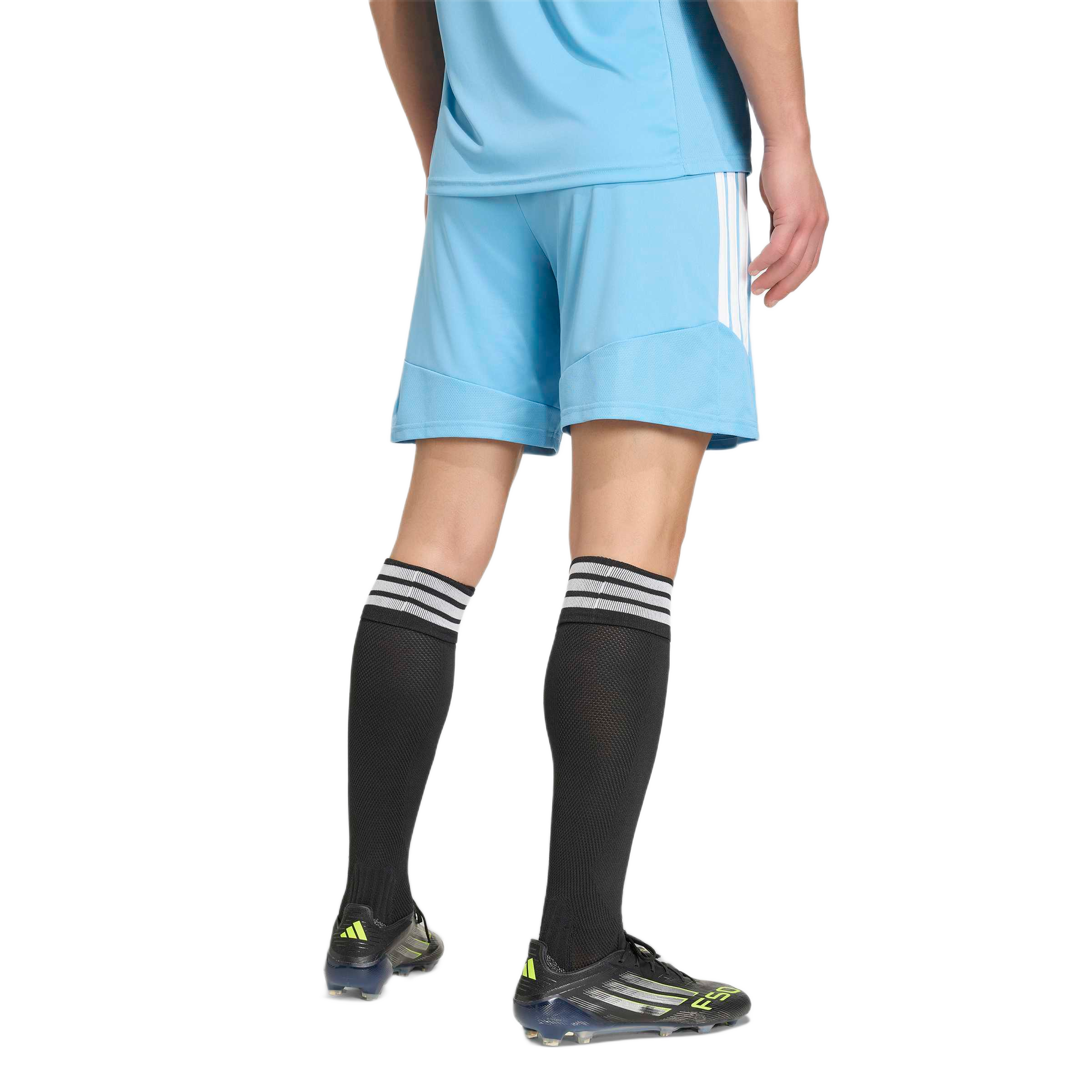 Rear view of a person wearing a light blue soccer kit, black knee-high socks with white stripes, and black soccer cleats with neon yellow accents against a grey background.