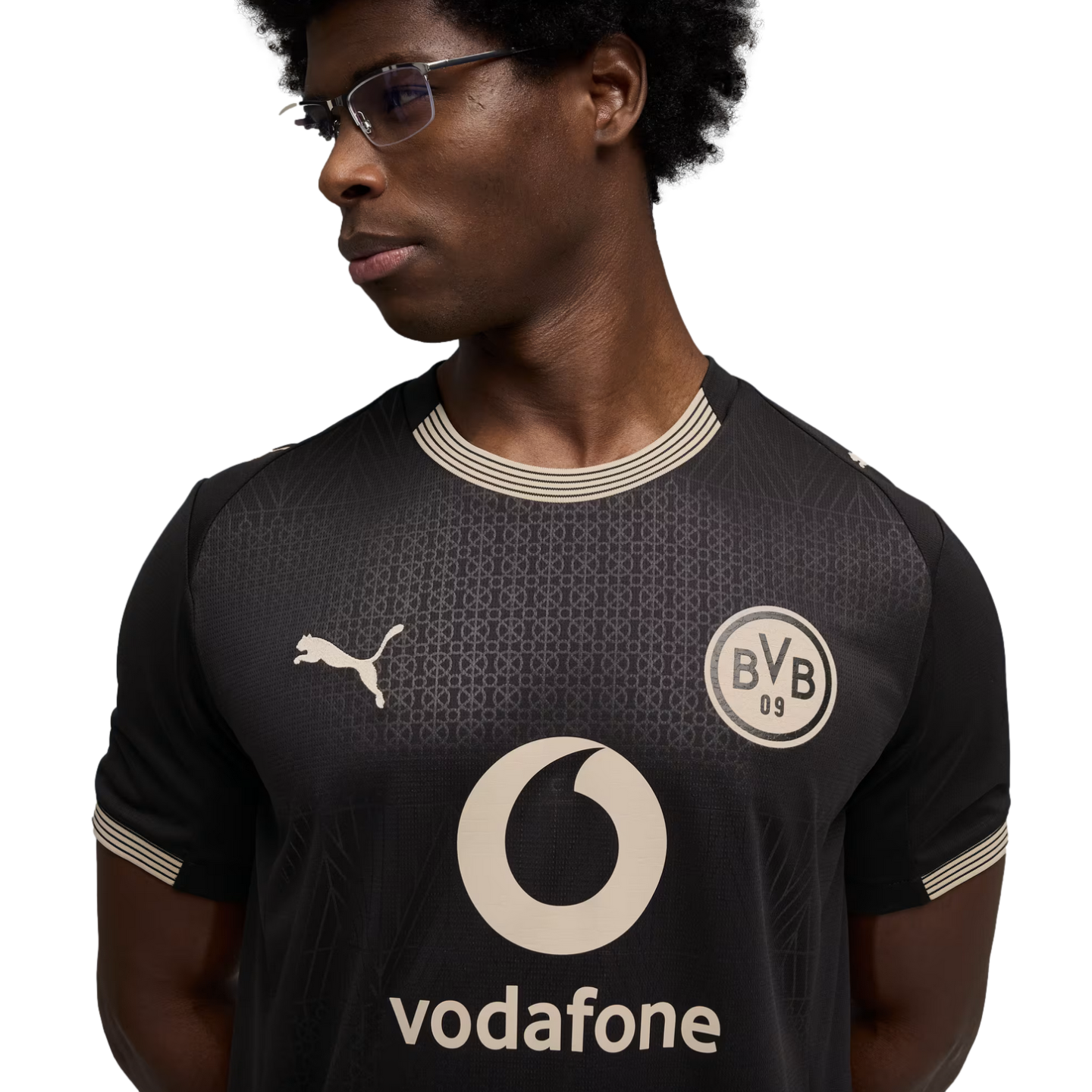 A man wearing glasses and a black Puma football jersey featuring the Borussia Dortmund (BVB) crest and a Vodafone sponsor logo, set against a black background.