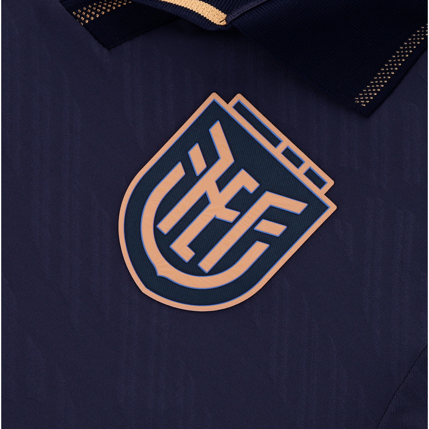 A close-up of a dark navy blue textured jersey featuring a stylized copper-colored crest in the center. The crest consists of interlocking geometric letters within a shield-like shape. A section of a navy blue ribbed collar with copper-toned stitching is visible at the top.