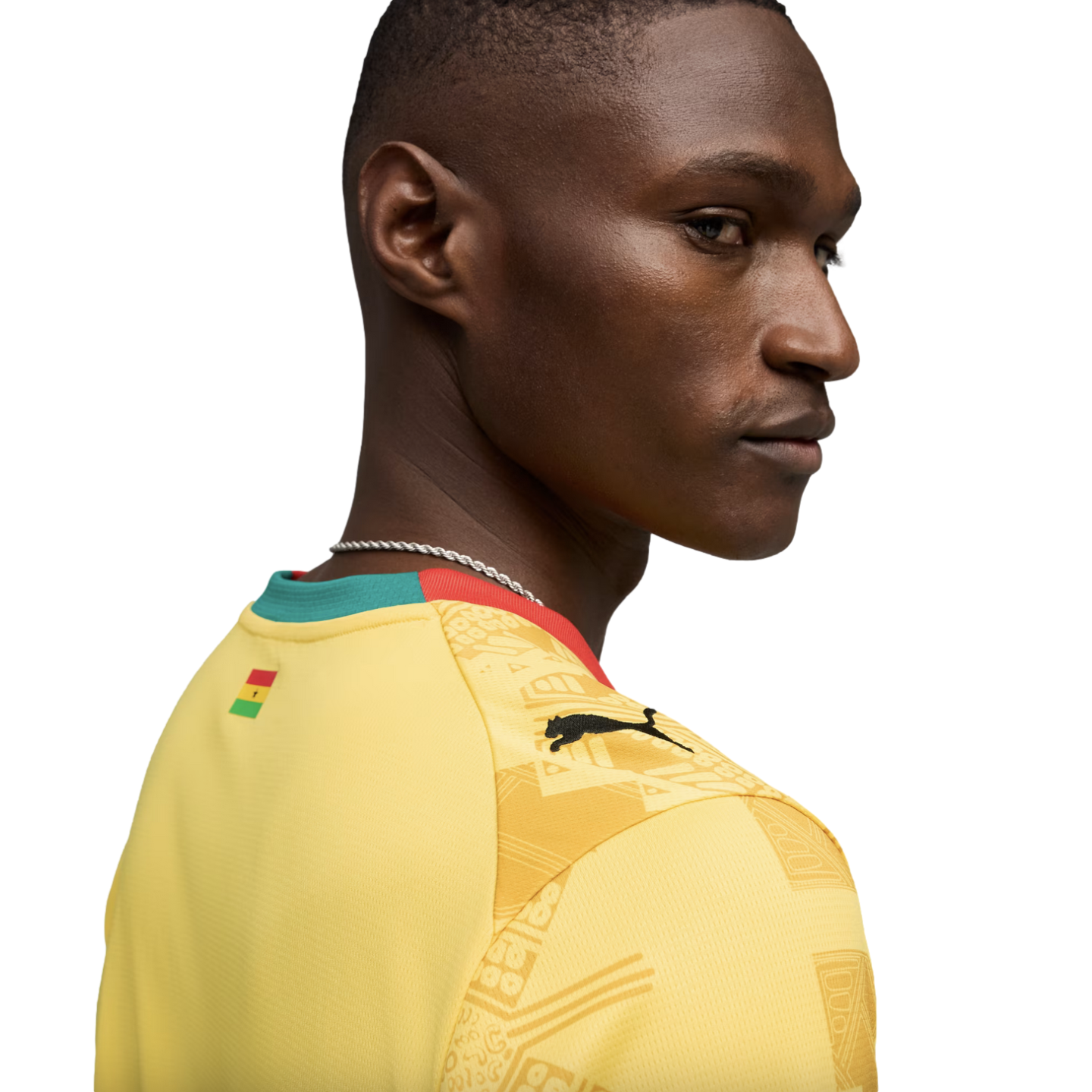A profile view of a Black man looking over his shoulder, wearing a yellow Puma sports jersey with the flag of Ghana on the back of the neck and a patterned design on the shoulder, set against a solid black background.