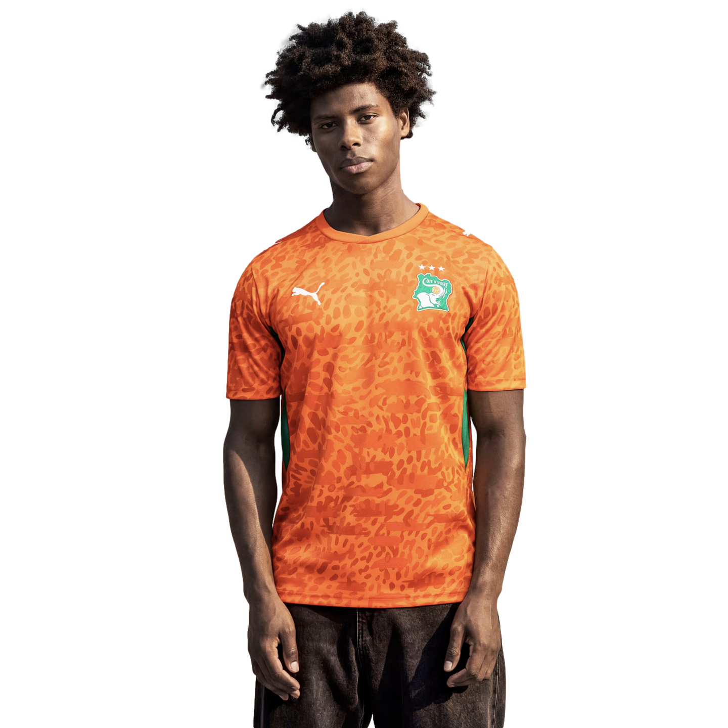 A medium shot of a young man with dark, curly hair wearing an orange Puma soccer jersey for the Côte d'Ivoire national team. The jersey features an all-over tonal leopard-print pattern, a green and white national crest on the left chest, and a white Puma logo on the right. He stands against a solid black background, looking directly at the camera while wearing dark-colored jeans.