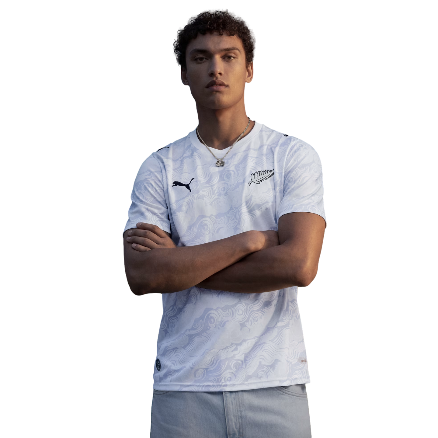 A waist-up portrait of a young man with curly hair posing with his arms crossed against a solid black background. He is wearing a white Puma jersey with a subtle, light-grey tonal swirl pattern. The jersey features a black Puma logo on the right chest and a black New Zealand silver fern emblem on the left chest. He is also wearing a thin silver necklace and light-colored pants.