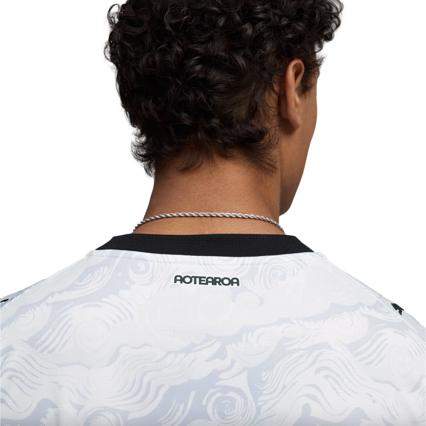 Close-up from behind of a person with dark, curly hair wearing a white jersey with a grey swirl pattern and a black collar. The word "AOTEAROA" is printed in black on the back of the jersey, just below the collar. A thin silver chain necklace is visible around the neck against a solid black background.