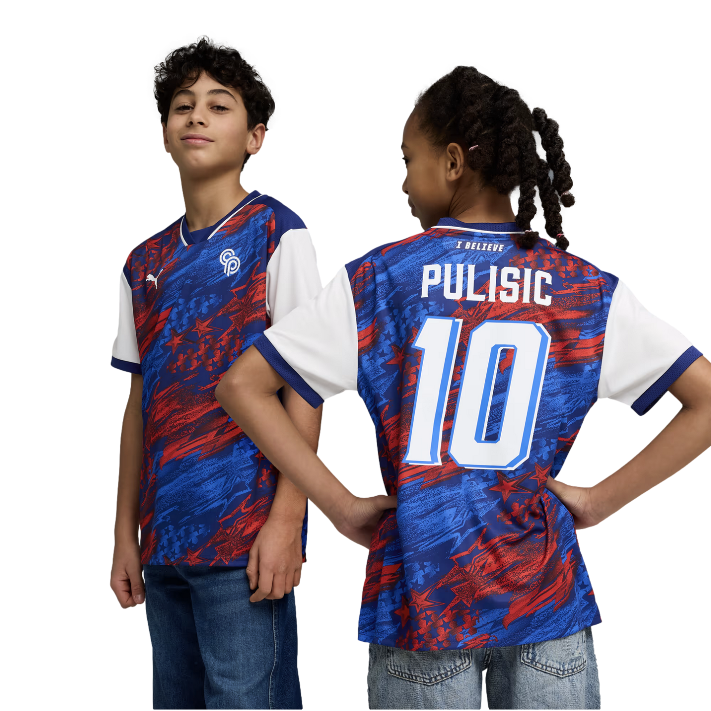 A studio portrait against a black background shows two young children modeling a blue and red Puma soccer jersey. The boy on the left faces forward, showing the front of the jersey with its abstract star pattern and Puma logo. The girl on the right faces away with her hands on her hips to show the back of the jersey, which features the name "PULISIC" and the number "10" in white.