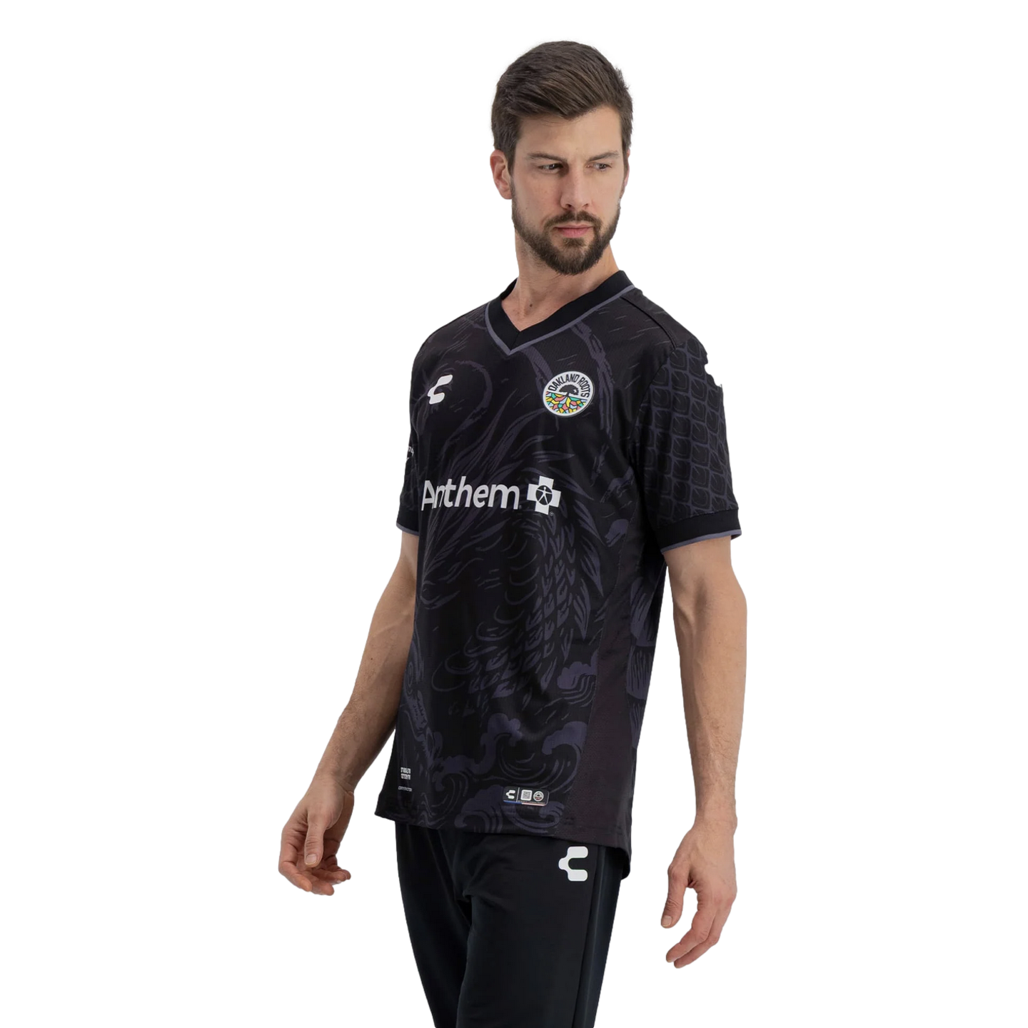 A man models an Oakland Roots SC soccer jersey against a solid black background. The black V-neck jersey features tonal grey wavy patterns on the front, textured scale-like patterns on the sleeves, the team's circular crest on the chest, and an Anthem sponsor logo. He is also wearing black athletic pants with a small white logo on the thigh.