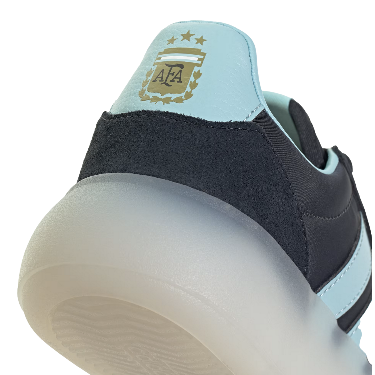 A close-up shot focusing on the heel of an Adidas shoe, featuring a light blue panel with the gold Argentina Football Association (AFA) crest and three stars. Below the blue panel is a dark navy textured material and a thick, semi-translucent white sole, set against a black background.