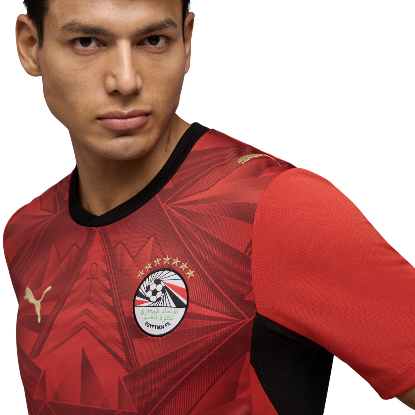 A close-up portrait of a man wearing a red Egypt national football team jersey by Puma. The jersey features an intricate geometric pattern, the Egyptian FA crest with seven gold stars, and gold Puma logos. He looks slightly off-camera against a solid black background.